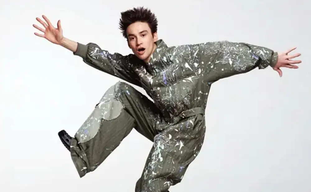 A young person in a paint-splattered jumpsuit jumps with arms outstretched against a white background.