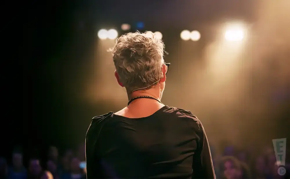 Back of a person with gray, curly hair on stage, facing a spotlight and an audience.