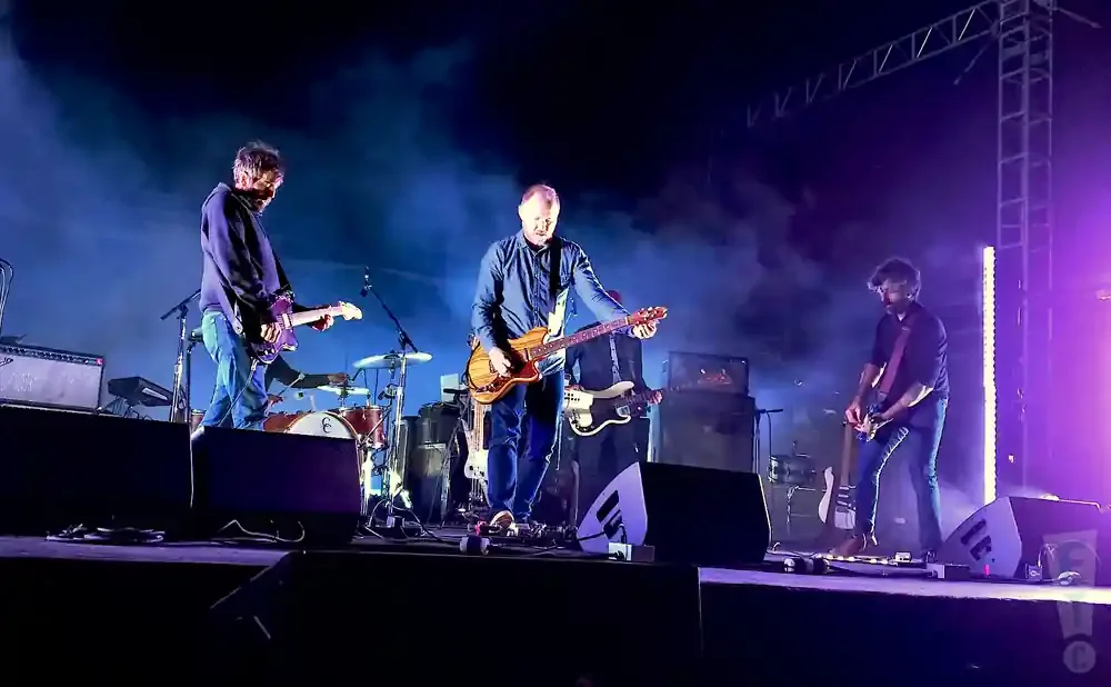 A band performs on a stage with purple lighting and fog.