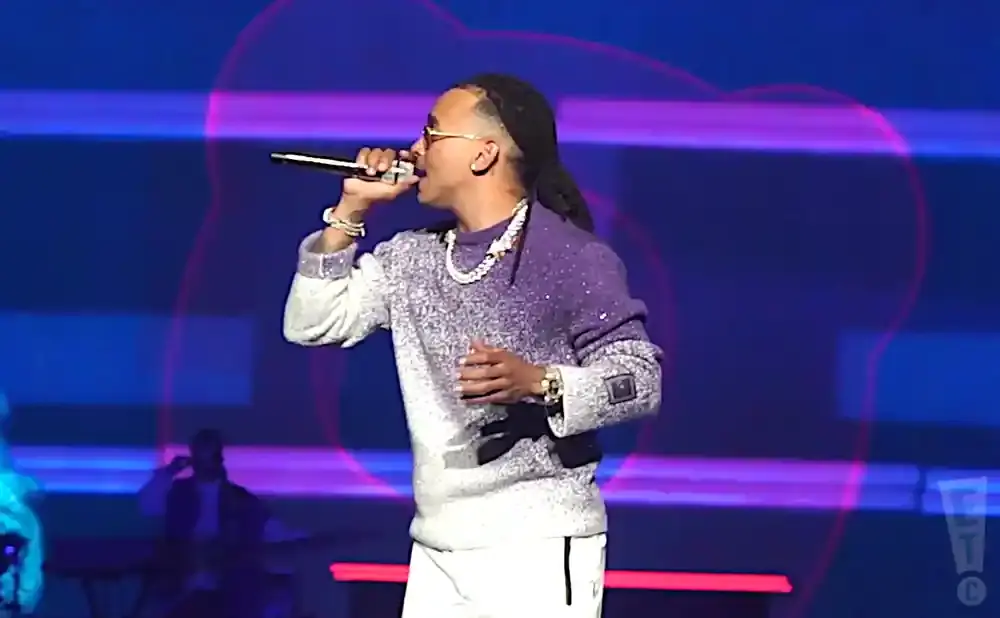 A male singer performs on stage, wearing a purple and white sparkly sweater and white pants, holding a microphone.