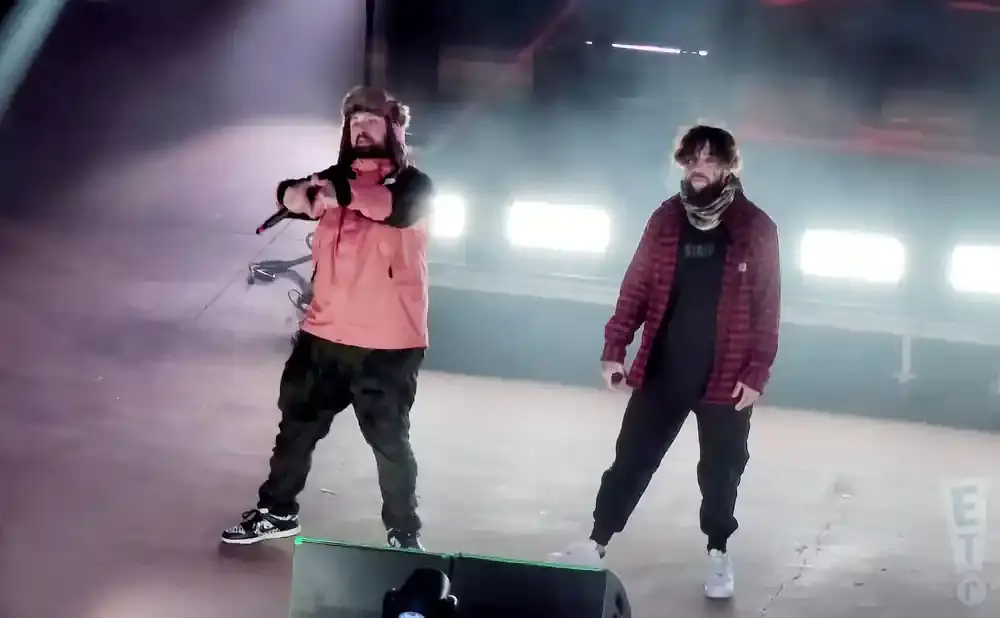 Two men perform on stage, one in an orange jacket and fur hat, the other in a red plaid shirt.