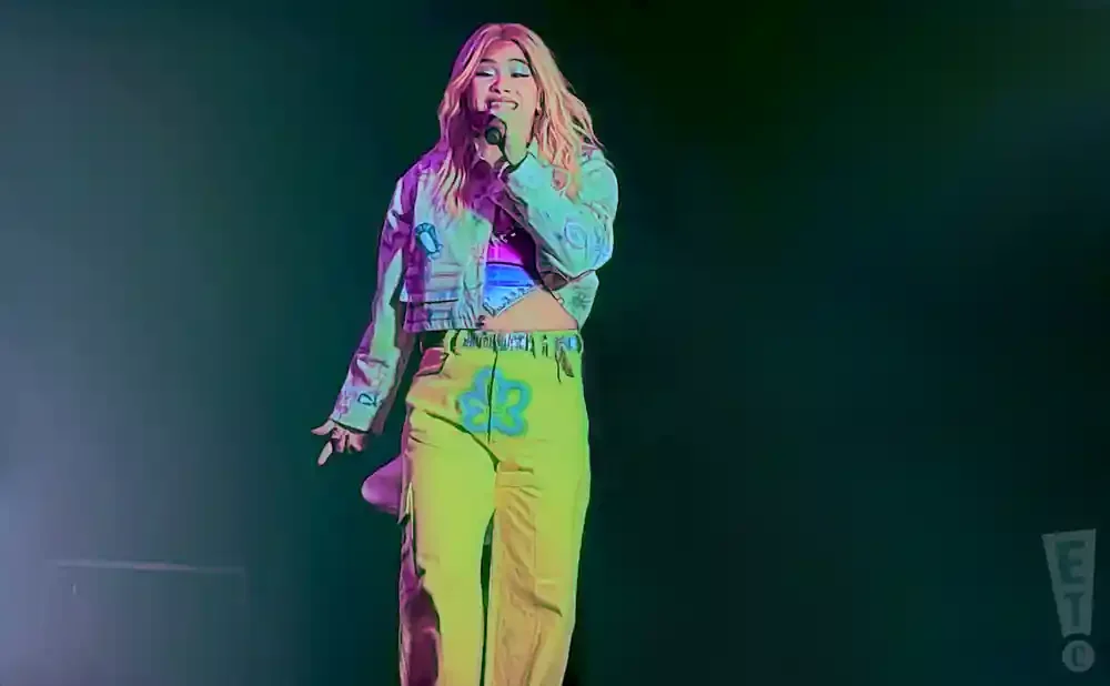 Singer performing on stage wearing a pink top, a decorated jacket, and bright yellow pants with a flower design.