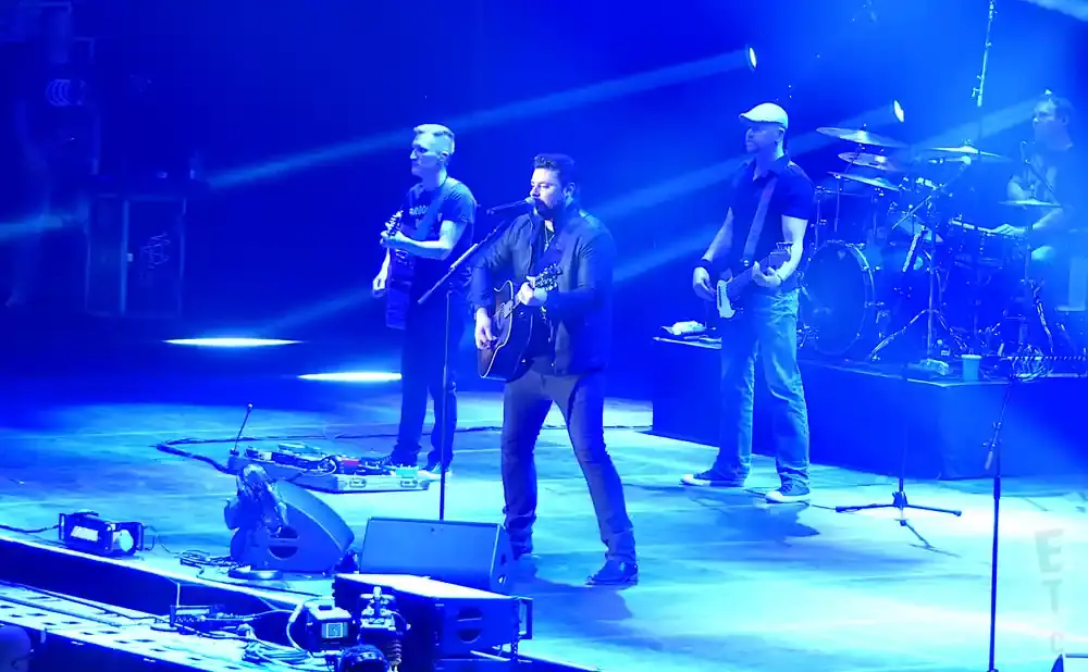 A band performs on a stage lit with blue spotlights, with singers playing guitars and a drummer in the background.