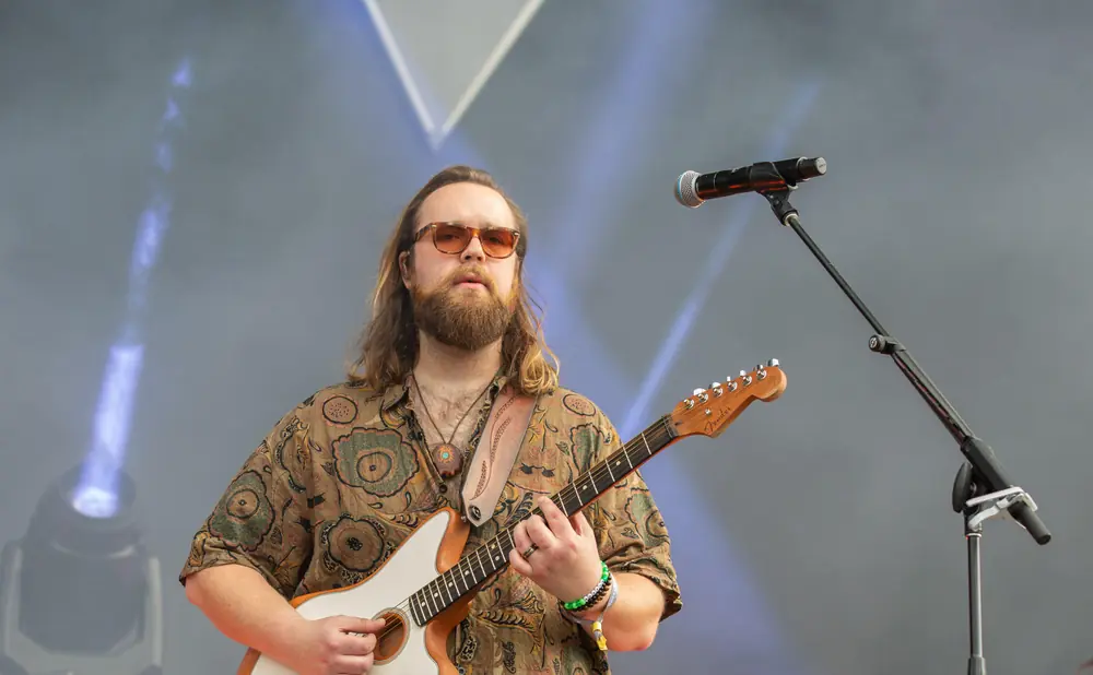 A male guitarist with long brown hair and sunglasses plays a white electric guitar on stage, with a microphone stand visible.