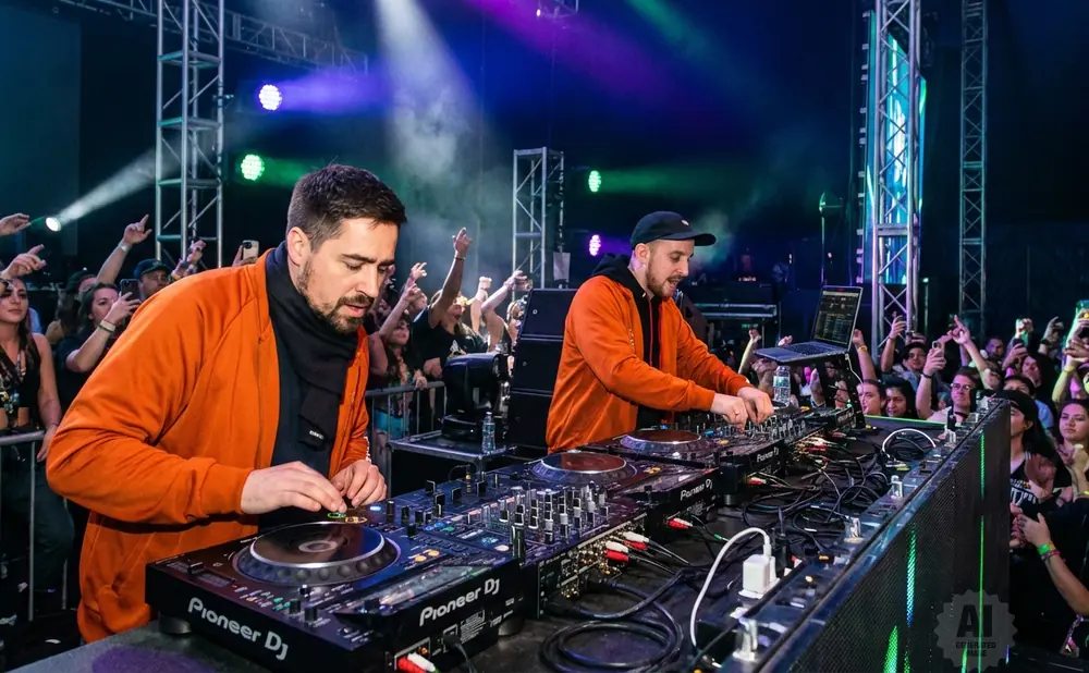 Two DJs in orange jackets perform at a concert with a cheering crowd.