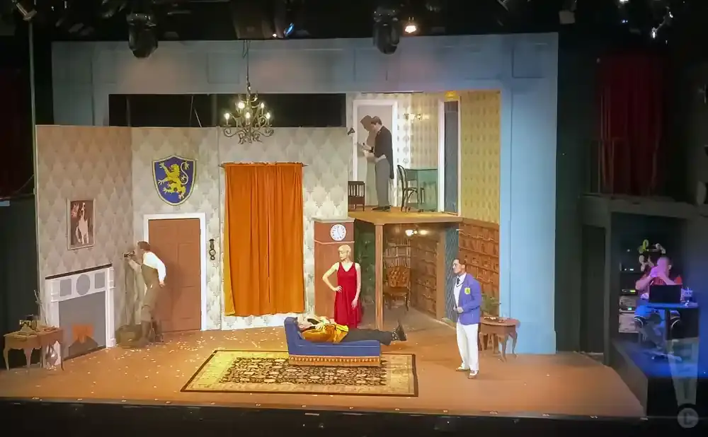 Stage set for a play with actors on multiple levels, depicting a living room and an upper floor.