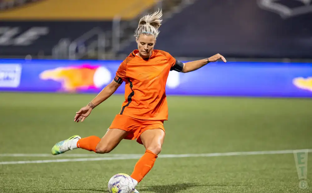 Woman in orange soccer uniform kicks a ball on a green field.