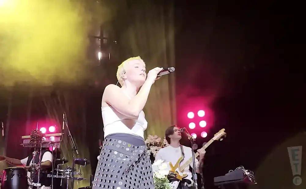 Singer with short blonde hair performs on stage with band members playing guitar and drums.