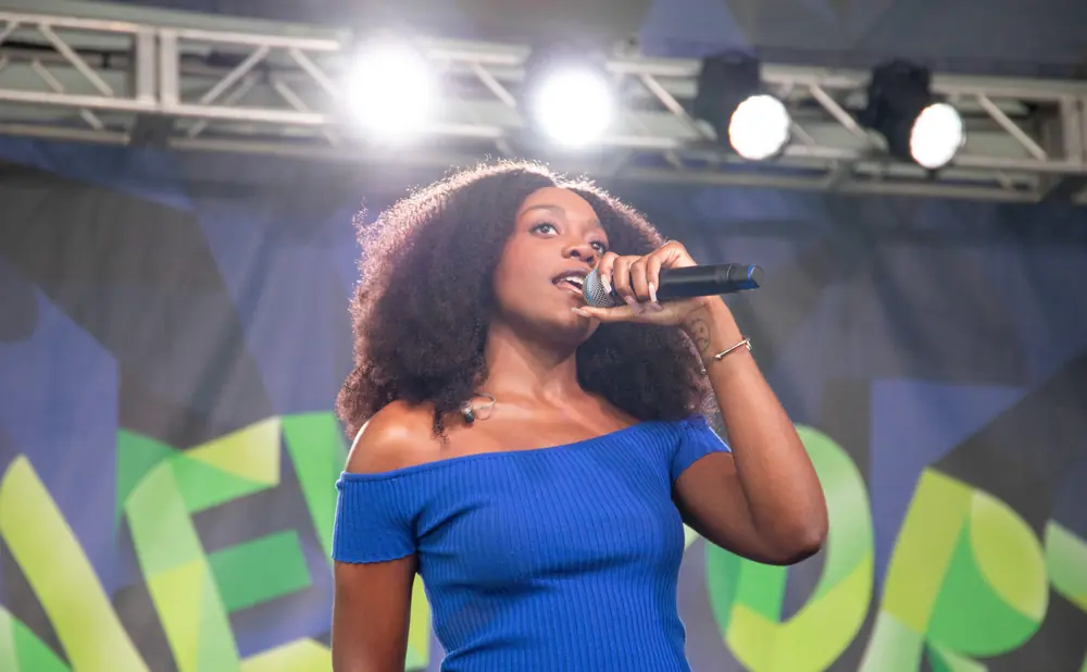 A woman with curly hair sings into a microphone on stage, wearing a blue off-the-shoulder top.