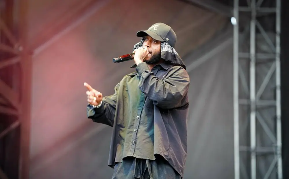 Rapper wearing a black hat and bandana, holding a microphone and gesturing on stage.