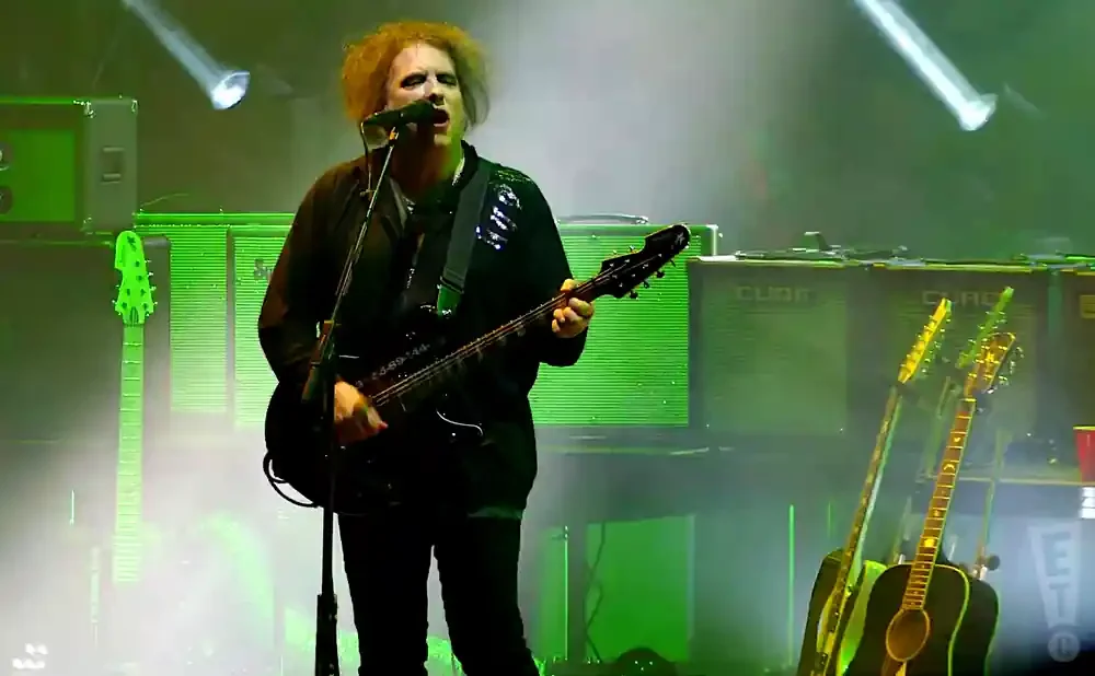 A man with curly hair sings and plays a black guitar on stage with green lighting.
