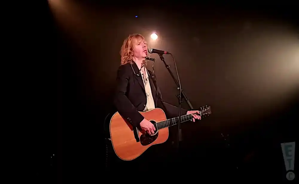 Musician with blonde hair plays an acoustic guitar and sings into a microphone while wearing a black jacket and a patterned shirt.