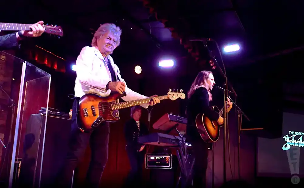 A band performs on stage. The lead singer, a white male with gray hair, plays a bass guitar. A woman with long hair sings into a microphone while playin…