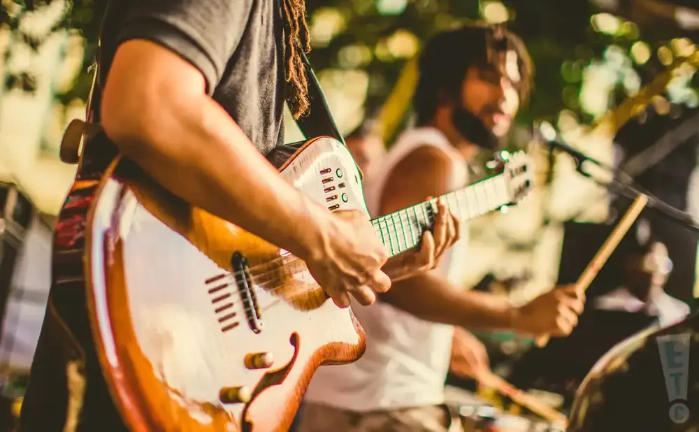 Musicians play guitars and drums outdoors, with a guitarist in the foreground playing a hollow-body electric guitar.