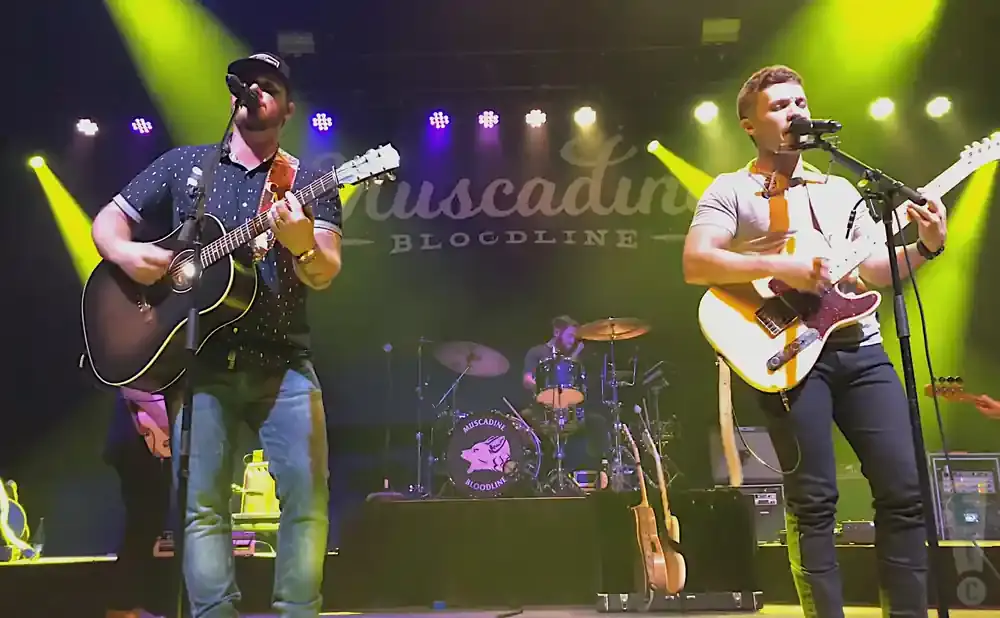 Two men perform on stage with guitars and microphones, with a drummer in the background.