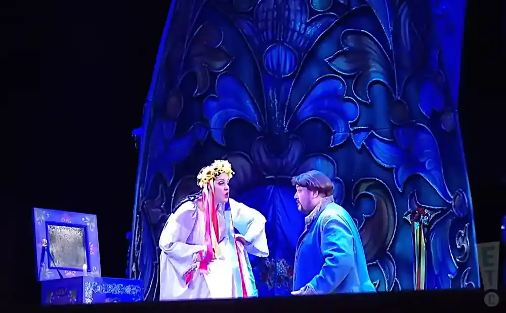 Stage actors in traditional costume on a blue ornate set. A woman wears a flower crown, a man wears a blue coat.