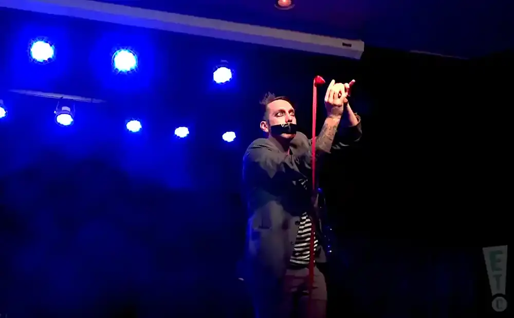 A man with black tape over his mouth stands on stage with his hands raised, holding a red ribbon.