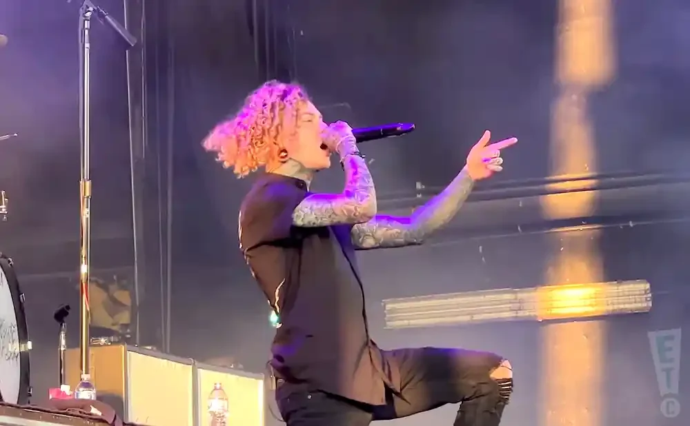 A tattooed singer with curly blonde hair sings into a microphone on stage, wearing a black shirt and ripped jeans.