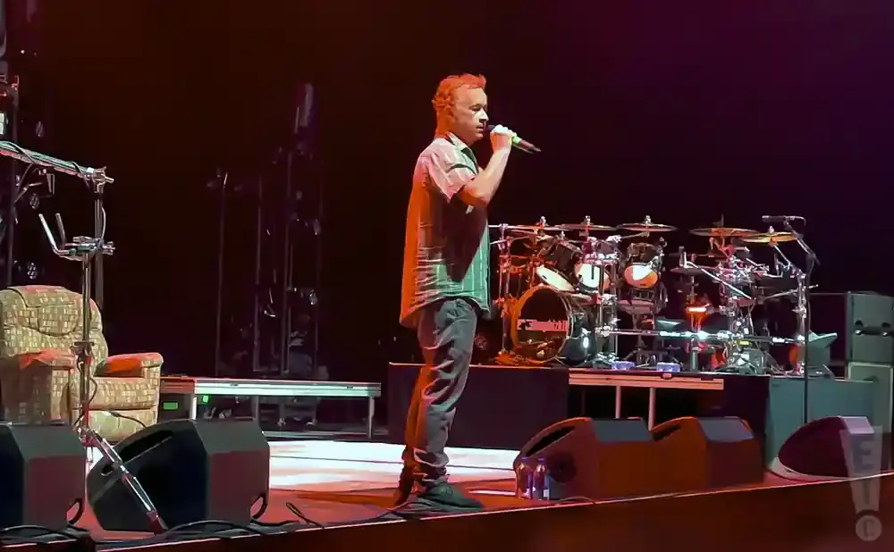 Musician singing into a microphone on a stage with a drum kit behind him.