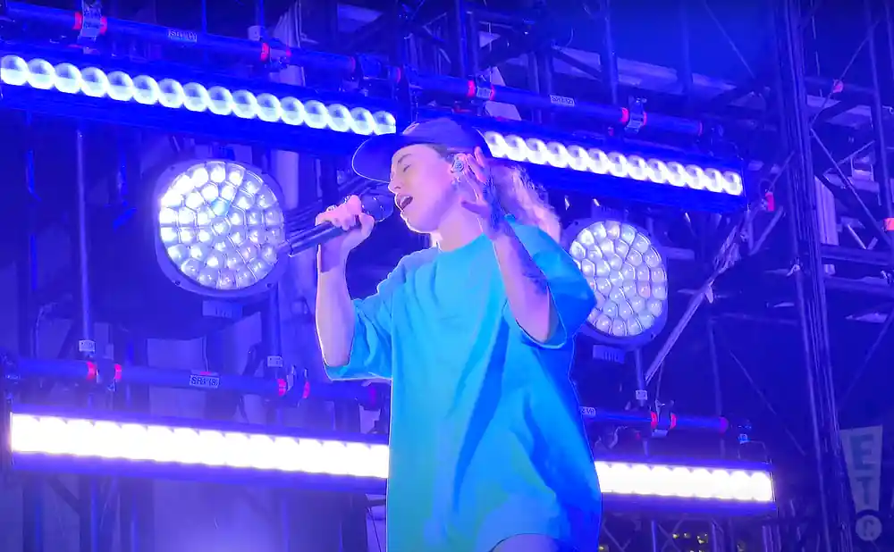 A singer in a blue t-shirt and cap performs on stage with bright blue lights.