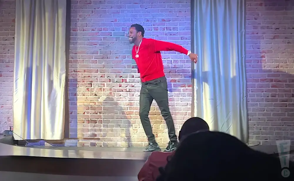 A man in a red sweater and dark pants stands on stage, smiling, with a brick wall and curtains behind him.