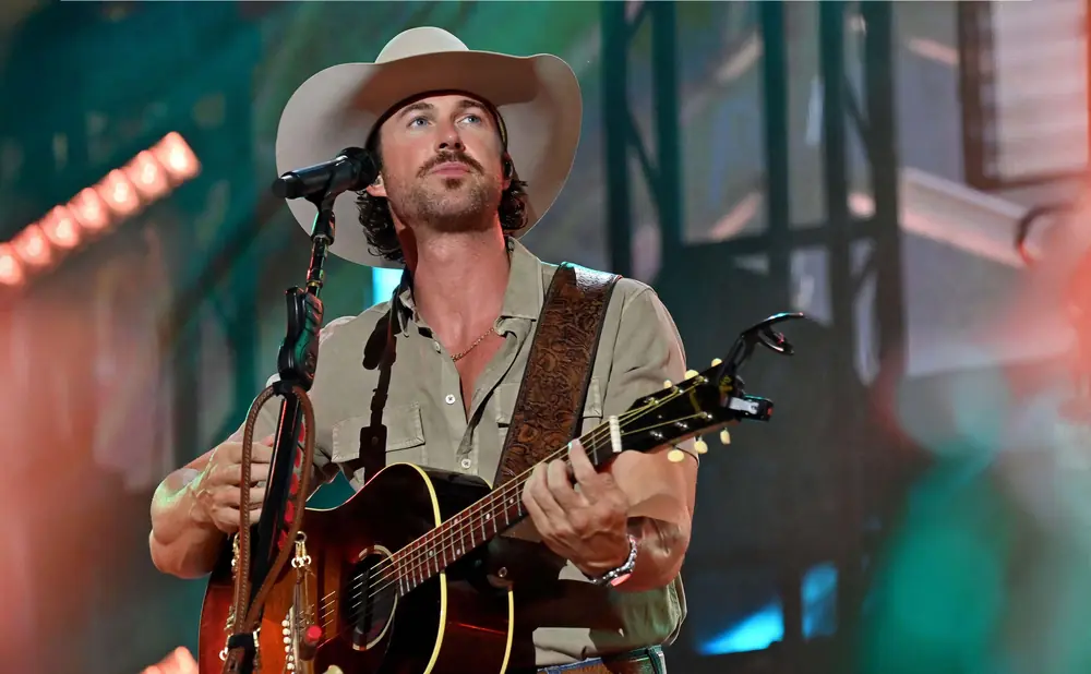 A man wearing a cowboy hat and a light brown shirt plays an acoustic guitar on stage.