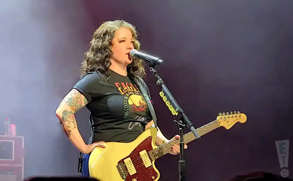 Singer with curly hair and tattoos plays a cream-colored electric guitar on stage.