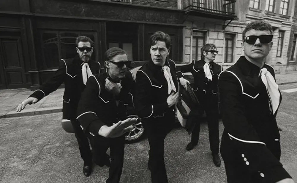 Five men in black suits and sunglasses stand on a street.
