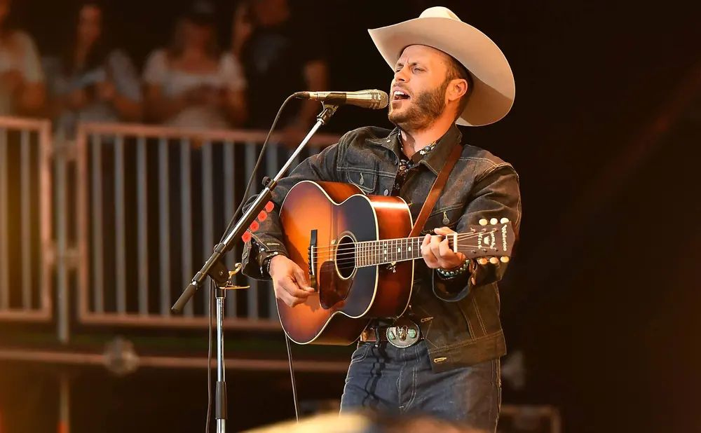 A man in a cowboy hat sings into a microphone while playing an acoustic guitar on stage.