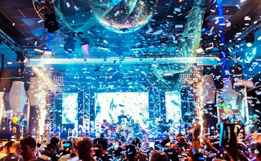 A DJ performs on a stage with confetti and fireworks at a crowded club, with a disco ball overhead.