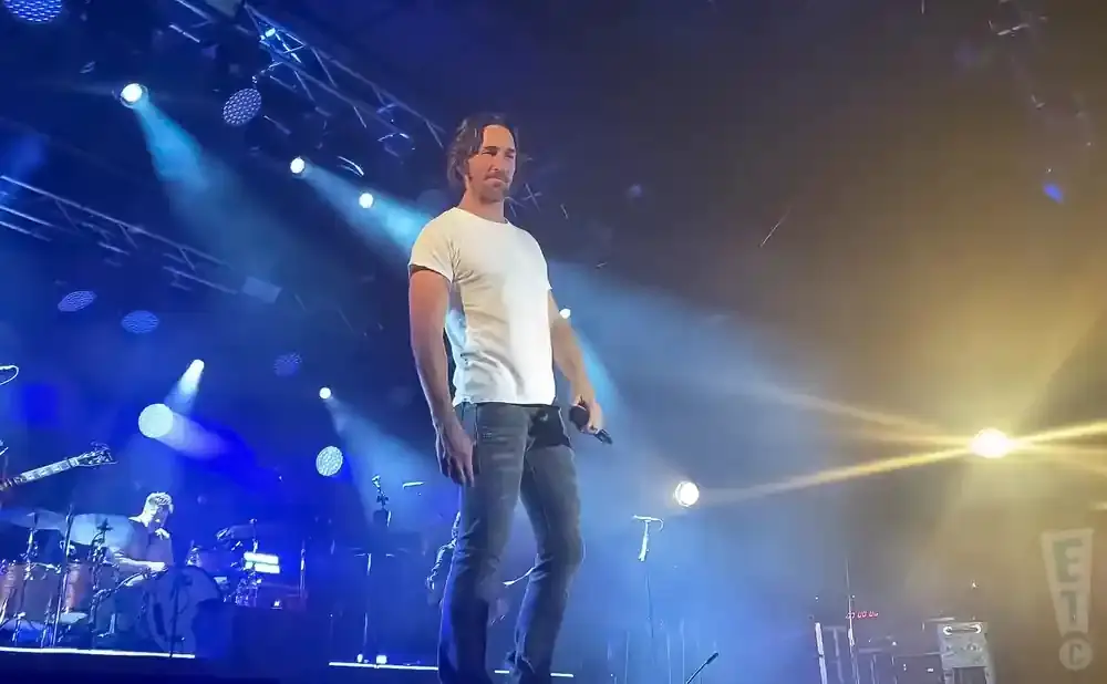 A man in a white t-shirt and jeans holds a microphone on a brightly lit stage.