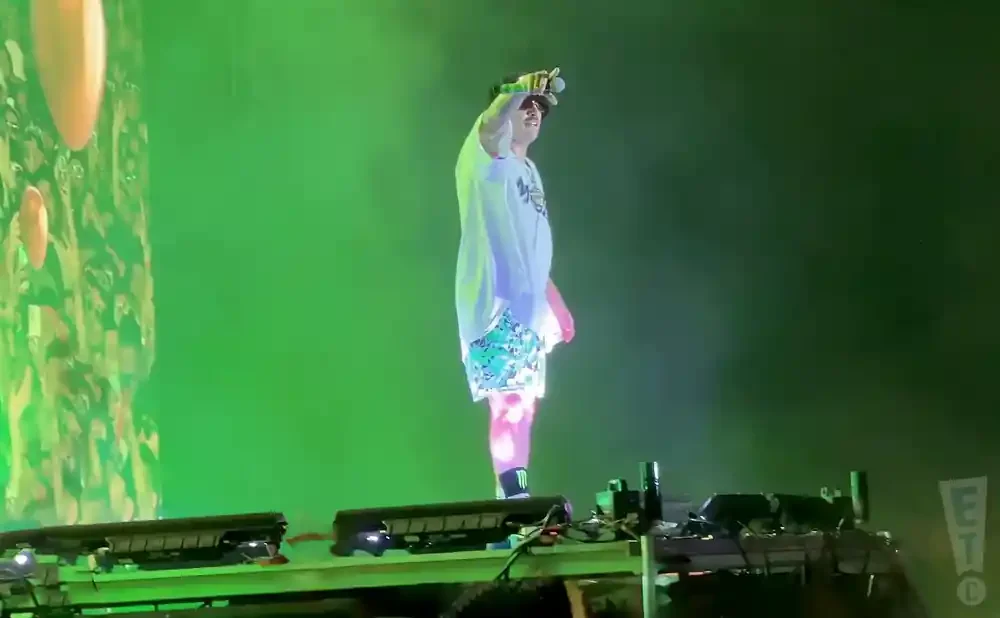 A performer in a white shirt and patterned shorts stands on stage, illuminated by green lights.