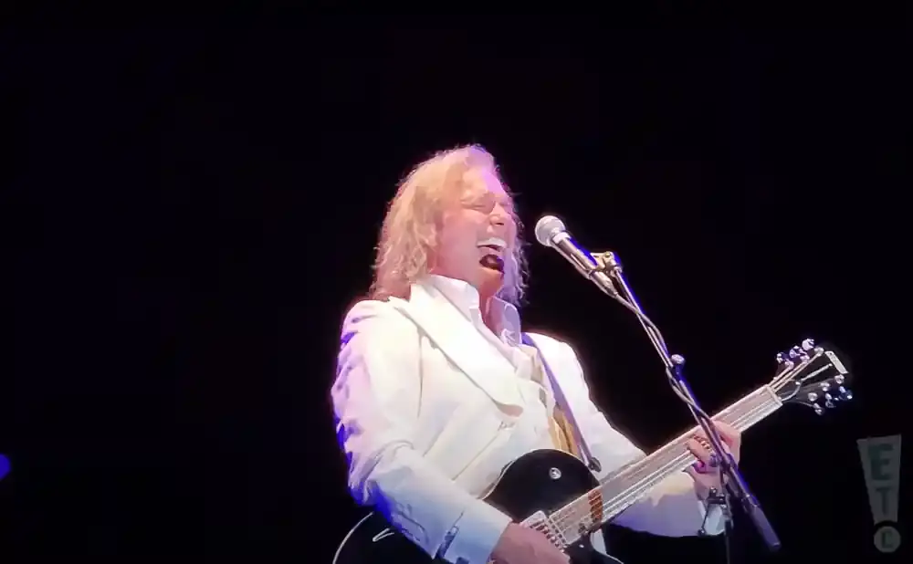 A male singer in a white suit plays a black electric guitar on stage.