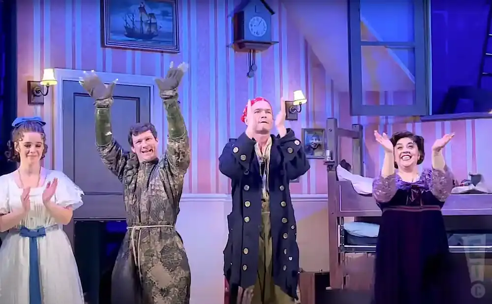 Four actors on a stage, two men and two women, clap with raised hands in front of a room with striped wallpaper.