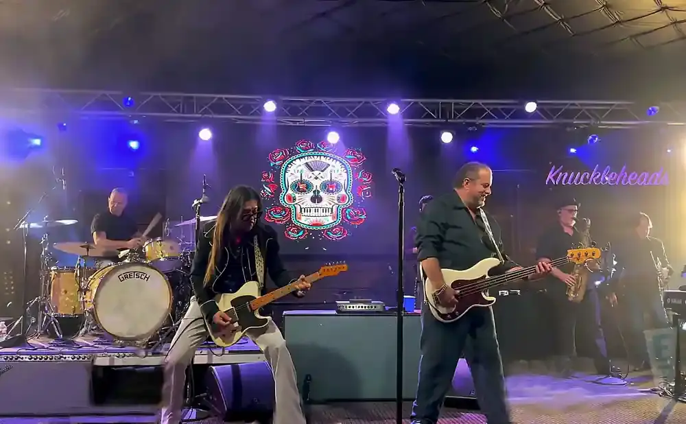 A rock band performs on stage with a colorful skull backdrop and neon 