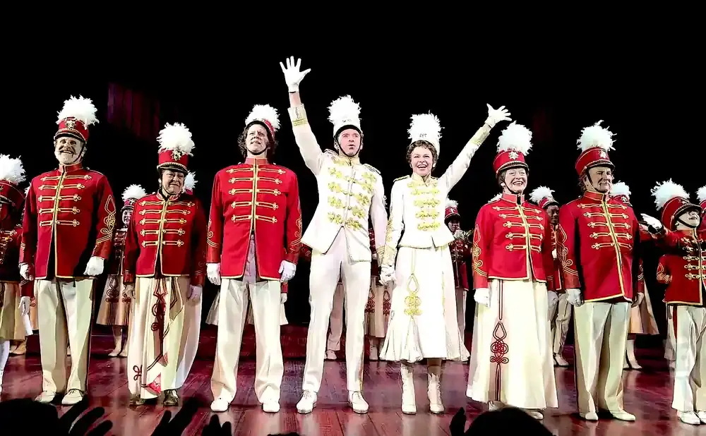 A cast in red and white marching band uniforms on stage, some waving.