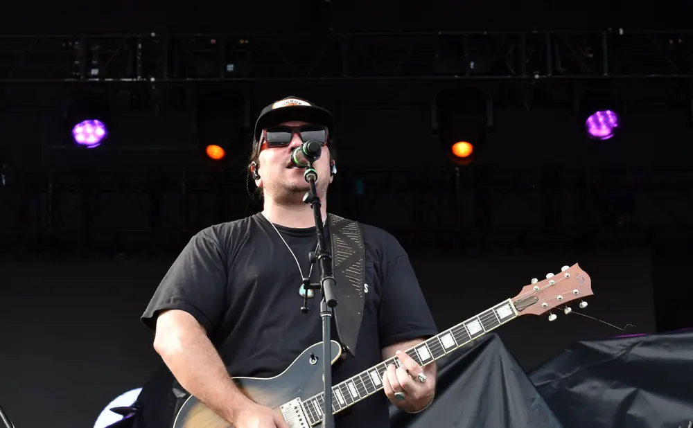 Musician wearing sunglasses and a black t-shirt plays a guitar on stage, with orange-patterned shorts visible.