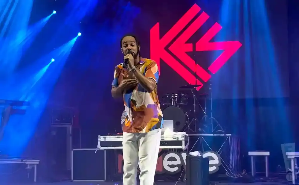Man in a colorful shirt sings into a microphone on a stage with blue lights and a large pink logo.