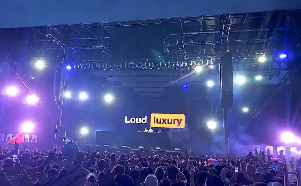 A crowd at an outdoor concert watches Loud Luxury perform on a brightly lit stage.