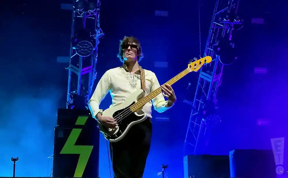 A male bassist plays a white Fender Precision bass on a stage illuminated by blue lights.