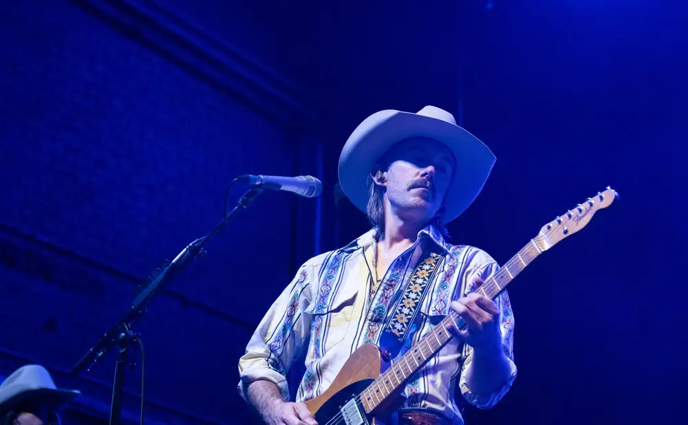 A man wearing a cowboy hat and a patterned shirt plays an electric guitar under blue stage lights.