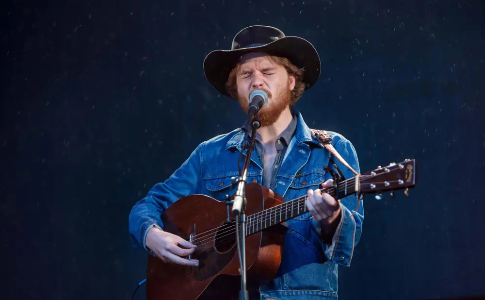 Musician in a cowboy hat and denim jacket plays an acoustic guitar and sings into a microphone.
