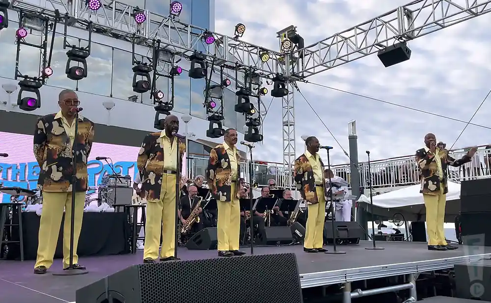 A group of male singers in matching patterned jackets and yellow pants perform on an outdoor stage with lighting rigs above.