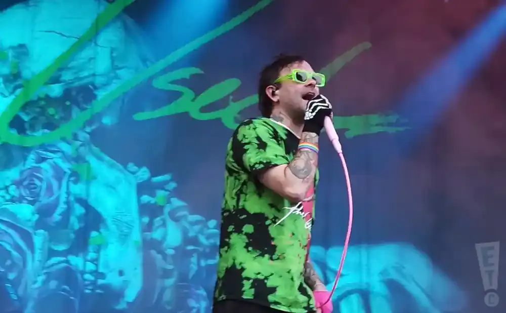 Man in a green and black tie-dye shirt and neon green glasses sings into a pink microphone on stage.