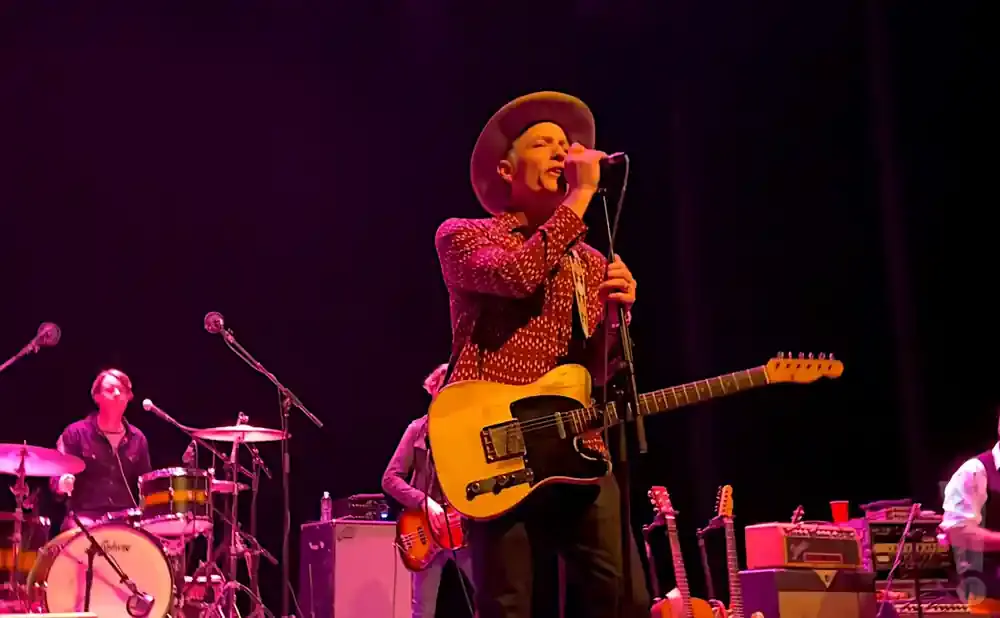A musician in a hat sings into a microphone while playing a guitar on stage.