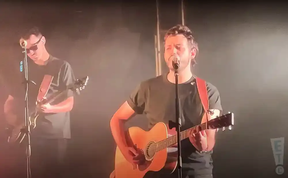 A band performs on stage with a singer in the foreground playing an acoustic guitar.