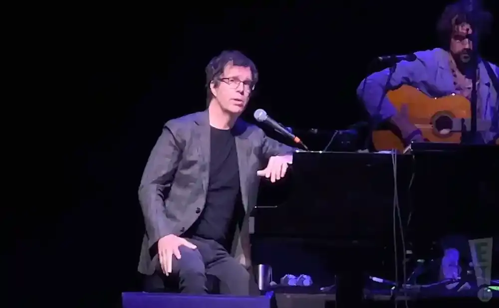 Musician sits at a piano, wearing glasses and a suit jacket, with a guitarist behind him playing an acoustic guitar.