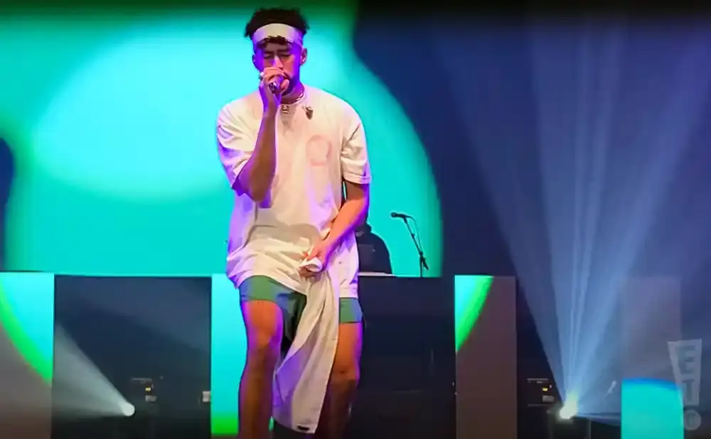 A male performer on stage in a white t-shirt, blue shorts, and a headband, holding a microphone and wiping his face with a white cloth.