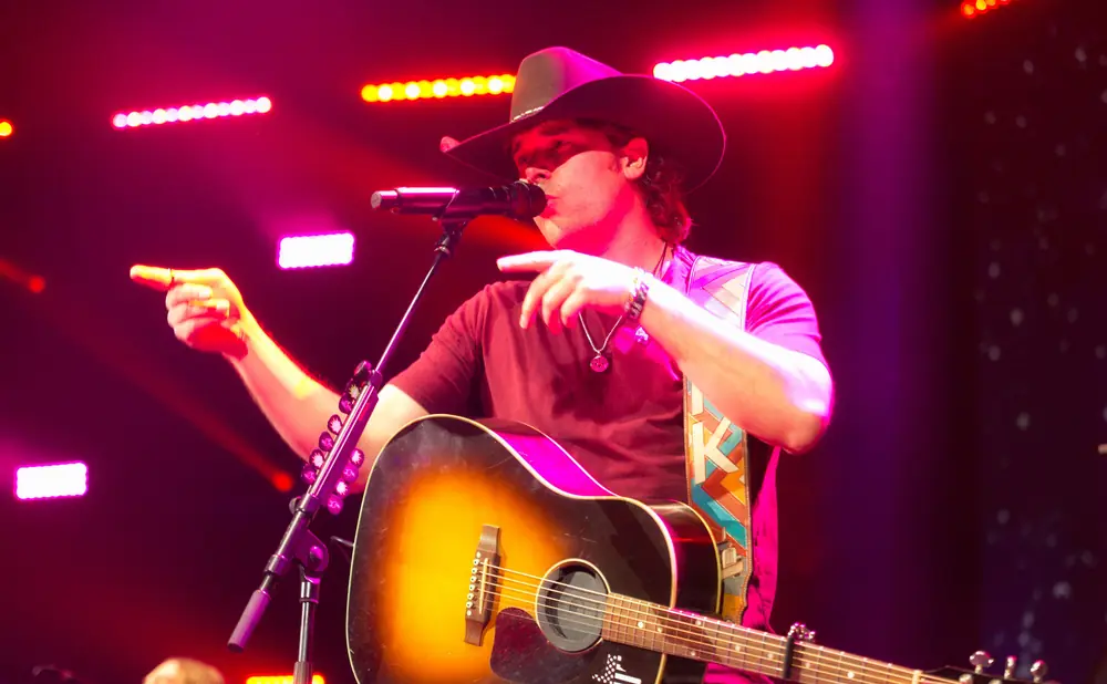 A country singer in a cowboy hat points while playing an acoustic guitar on a stage lit with pink and red lights.