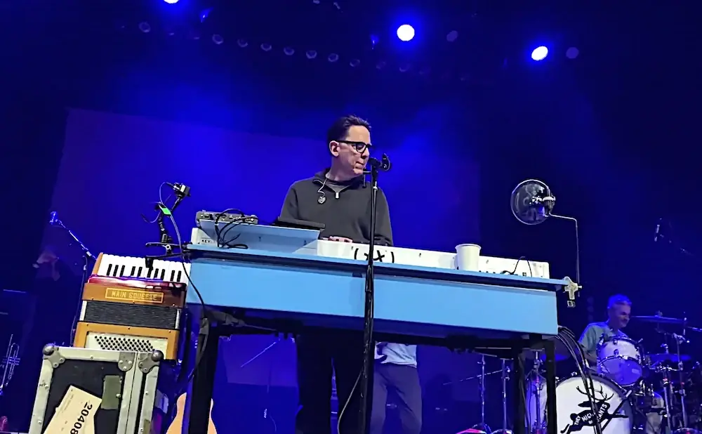 A man in glasses plays a keyboard on stage, with a drummer in the background.